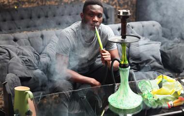 A young African American sits and smokes a hookah in a hookah room. Portrait of a man on a dark background.Smoke from a hookah.